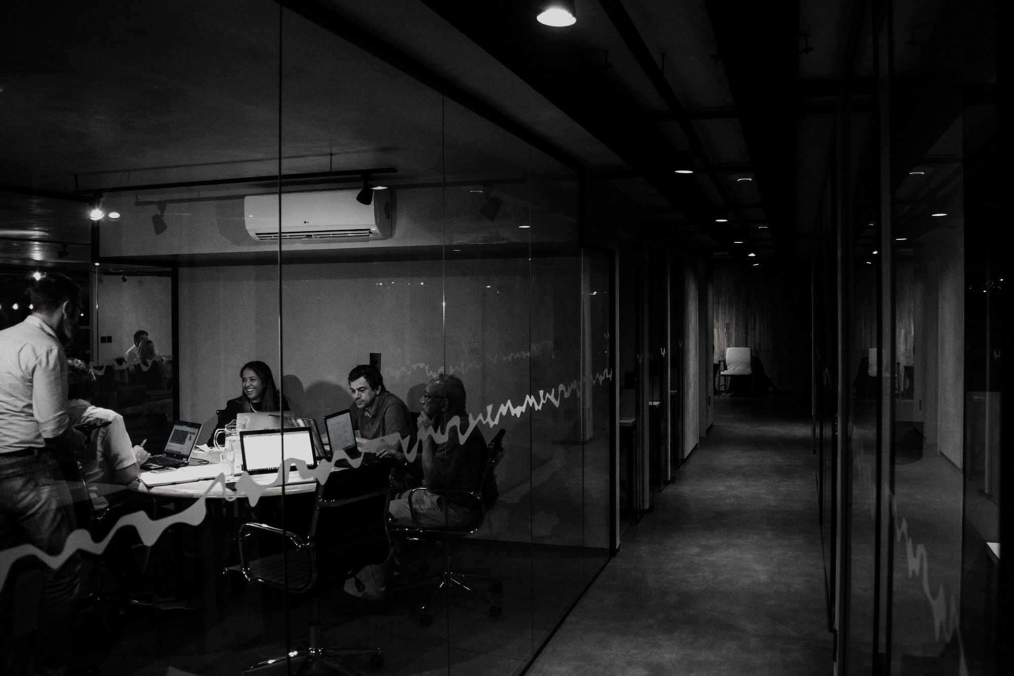 A team working late around a conference table, a data waveform tracing across the glass wall